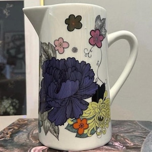 May include: White ceramic pitcher with a curved handle and spout. The pitcher is decorated with a large, detailed purple flower, yellow and orange flowers, and smaller pink and brown floral accents. The design is against a white background.