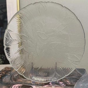 May include: A clear, round glass plate with an etched floral design. The plate features a scalloped edge and is supported by a clear stand. The etched design includes stylized leaves and flowing lines, creating a delicate, artistic appearance.