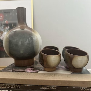 May include: A ceramic vase and four small bowls. The vase is round with a long neck, and the bowls are spherical. All pieces have a brown, gray, and white color scheme and sit on wooden bases. The items are displayed on top of books.