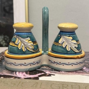 May include: A ceramic salt and pepper shaker set with a handle. The shakers are teal with yellow and blue accents, and a leaf design. The base is off-white with a blue wave pattern. The handle is teal.