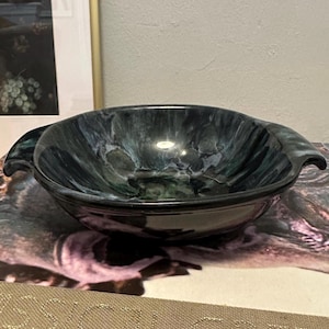 May include: A dark, marbled ceramic bowl with two handles. The bowl has a glossy finish and a swirling pattern of dark blue, green, and black. The bowl is likely used for serving food or as a decorative piece.