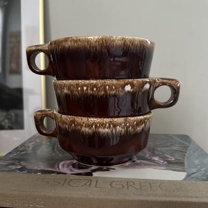 May include: A stack of three ceramic soup bowls with handles, featuring a deep brown glossy glaze and a distinctive white drip pattern along the upper rim. Each bowl has a wide, circular opening and a sturdy side handle for easy gripping.