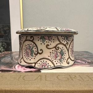May include: A decorative ceramic trinket box with a lid. The box is off-white with a floral pattern in pink and green, accented by brown swirls. The lid mirrors the design. The box is likely used for storing small items.