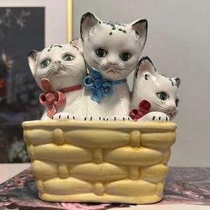 May include: A ceramic figurine featuring three white cats with green eyes and black-tipped ears, each wearing a bow. They sit in a yellow basket-shaped container. The cats have delicate painted details and are arranged closely together.