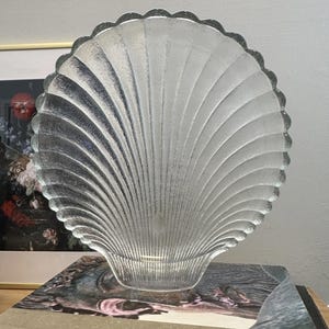 May include: A clear, glass seashell-shaped decorative dish. The dish has a scalloped edge and a textured, fan-like design. The dish is sitting on top of a book.