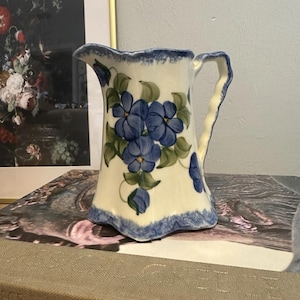 May include: A white ceramic pitcher with a blue floral design and a blue rim. The pitcher has a curved handle and a flared base. The flowers are a deep blue with green leaves. The pitcher is likely used for serving beverages.