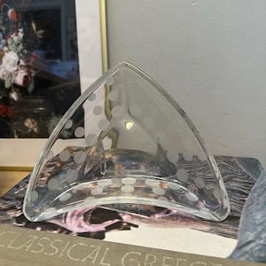 May include: A clear glass, triangular-shaped dish with etched polka dots. The dish has a curved base and is resting on a stack of books. A framed painting is in the background.