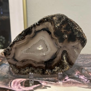 May include: A polished agate geode slice, showcasing concentric bands of color. The center features a white crystalline area, surrounded by layers of light purple, brown, and black. The geode is displayed on a clear acrylic stand.