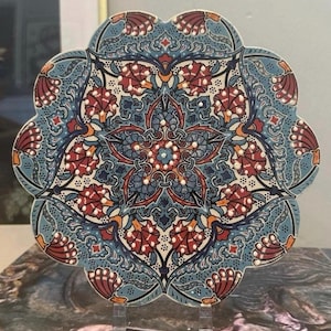 May include: A decorative ceramic coaster with a floral design. The coaster features a blue background with red and white floral patterns. The coaster is round with a scalloped edge and is displayed on a clear stand.