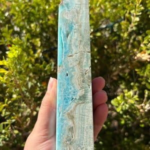 May include: A rectangular, polished crystal obelisk with a pointed top. The crystal features a blend of light blue, white, and tan colors with natural veining and patterns. The obelisk is held in a hand against a backdrop of green foliage.