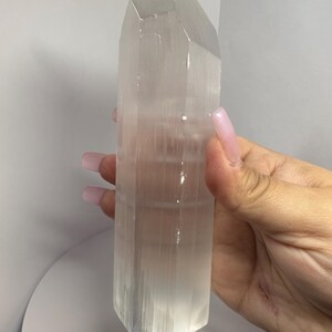 May include: A clear, rectangular selenite crystal tower with a pointed top. The crystal is held in a hand and has a translucent appearance. The tower is a light, off-white color and has vertical striations.