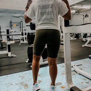 May include: A white t-shirt with the text "Nobody Cares Work Harder" printed on the back. The person wearing the shirt is standing in a gym.