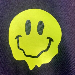 Puede incluir: Una tela azul marino con un gran gr&aacute;fico de carita sonriente de color amarillo ne&oacute;n. La carita sonriente tiene un dise&ntilde;o derretido y ojos y boca negros. La tela tiene un aspecto texturizado.