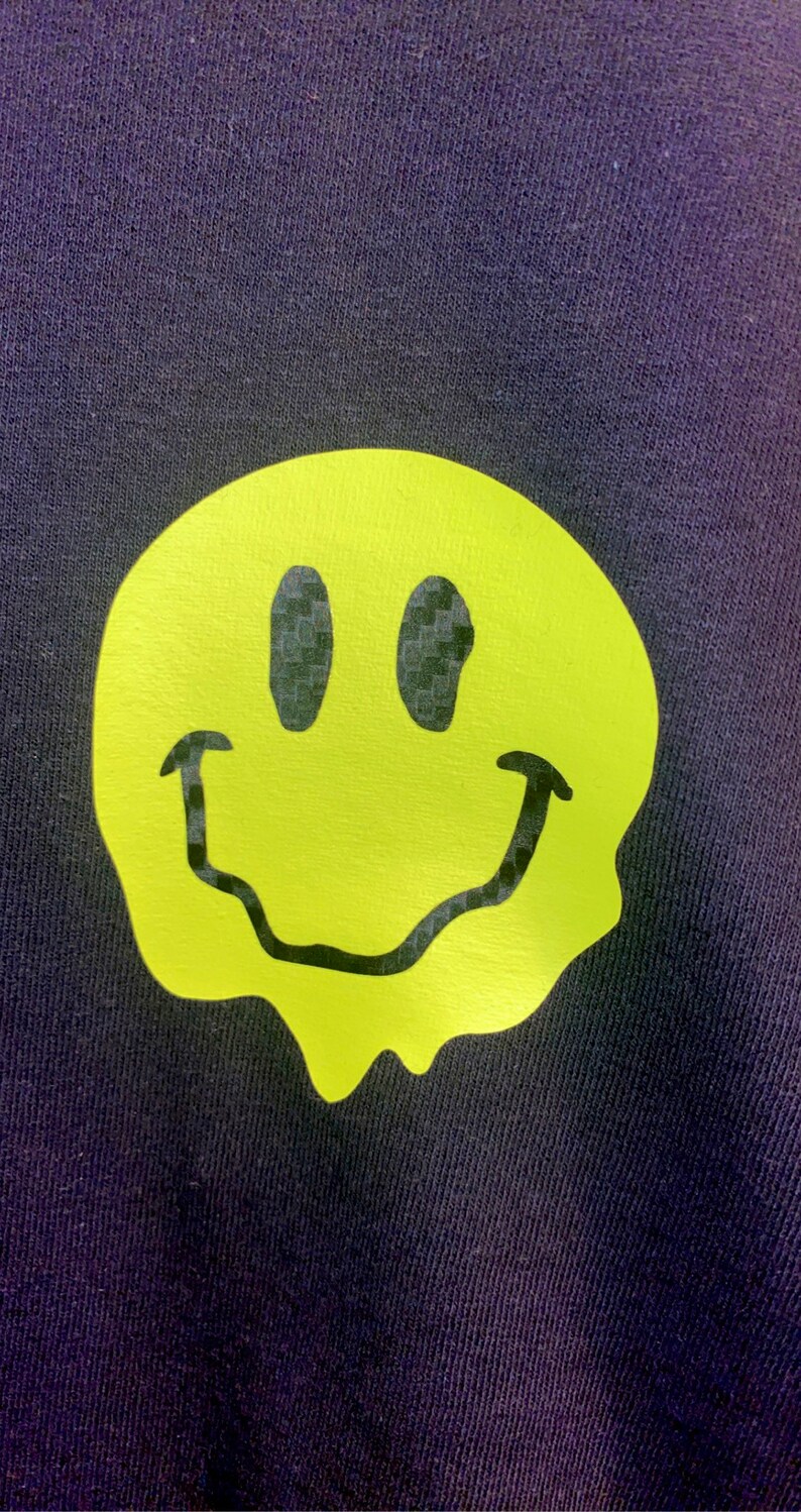 Puede incluir: Una tela azul marino con un gran gr&aacute;fico de carita sonriente de color amarillo ne&oacute;n. La carita sonriente tiene un dise&ntilde;o derretido y ojos y boca negros. La tela tiene un aspecto texturizado.