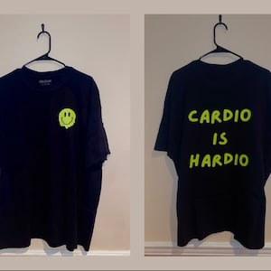 Puede incluir: Camiseta negra con un gr&aacute;fico de carita sonriente verde ne&oacute;n derriti&eacute;ndose en la parte delantera. La parte trasera de la camiseta presenta las palabras "CARDIO IS HARDIO" en verde ne&oacute;n. La camiseta est&aacute; colgada de una percha negra.
