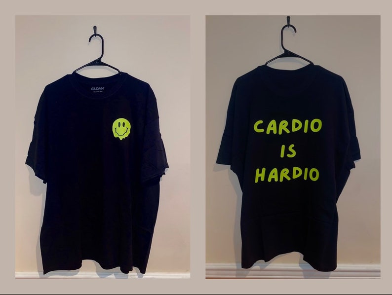 Puede incluir: Camiseta negra con un gr&aacute;fico de carita sonriente verde ne&oacute;n derriti&eacute;ndose en la parte delantera. La parte trasera de la camiseta presenta las palabras "CARDIO IS HARDIO" en verde ne&oacute;n. La camiseta est&aacute; colgada de una percha negra.