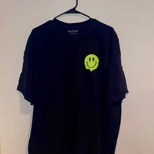 Puede incluir: Camiseta negra Gildan con un gr&aacute;fico de cara sonriente derriti&eacute;ndose en verde ne&oacute;n en la parte delantera. La camiseta est&aacute; colgada de una percha negra contra una pared blanca. El logotipo de Gildan es visible en el cuello.
