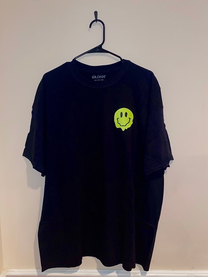 Puede incluir: Camiseta negra Gildan con un gr&aacute;fico de cara sonriente derriti&eacute;ndose en verde ne&oacute;n en la parte delantera. La camiseta est&aacute; colgada de una percha negra contra una pared blanca. El logotipo de Gildan es visible en el cuello.