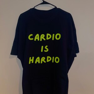 Puede incluir: Camiseta negra con las palabras "CARDIO IS HARDIO" en letras verde brillante. La camiseta est&aacute; colgada en una percha negra contra una pared neutra. El texto est&aacute; centrado en la parte posterior de la camiseta.