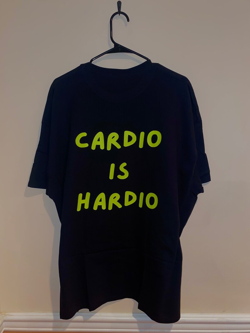Puede incluir: Camiseta negra con las palabras "CARDIO IS HARDIO" en letras verde brillante. La camiseta est&aacute; colgada en una percha negra contra una pared neutra. El texto est&aacute; centrado en la parte posterior de la camiseta.