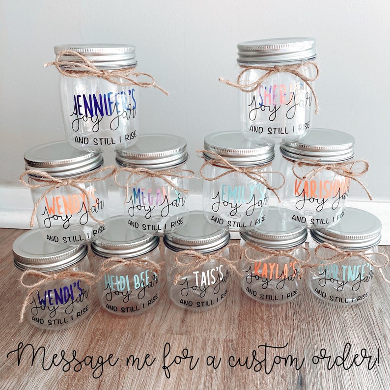 Pu&ograve; includere: Una collezione di barattoli di vetro trasparente con coperchi argentati e fiocchi di spago. Ogni barattolo ha un nome personalizzato e il testo "Joy Jar" e "And Still I Rise".