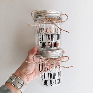 Pu&ograve; includere: Due barattoli di vetro trasparente con coperchi argentati e spago legato attorno alla parte superiore. I barattoli sono decorati con lettere in vinile nero che dicono "Amelia's 1st Trip to the Beach" e "Elliot's 1st Trip to the Beach".