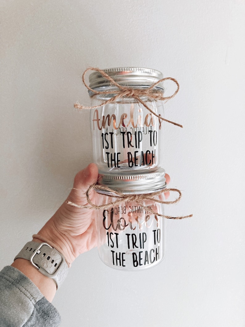 Pu&ograve; includere: Due barattoli di vetro trasparente con coperchi argentati e spago legato attorno alla parte superiore. I barattoli sono decorati con lettere in vinile nero che dicono "Amelia's 1st Trip to the Beach" e "Elliot's 1st Trip to the Beach".
