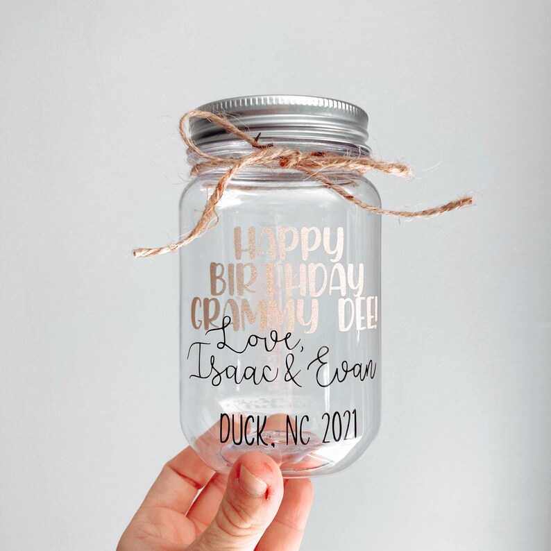 Pu&ograve; includere: Un barattolo di vetro trasparente con un coperchio argentato e un fiocco di spago marrone. Il barattolo ha un adesivo in vinile color oro rosa che dice "Happy Birthday Grammy Dee! Love, Isaac & Evan Duck, NC 2021".