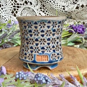 May include: A cylindrical ceramic planter with a flared rim and a footed base. The exterior features a repeating pattern of dark blue flowers and triangles on a cream-colored background. A rectangular stamp with embossed characters is visible.