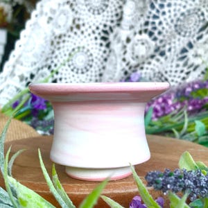 May include: A small, handcrafted ceramic vessel with a flared rim and base. The piece features a soft pink and white marbled design. The vessel is displayed on a wooden surface, surrounded by greenery and purple flowers.