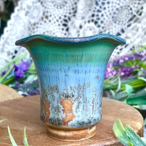 May include: A handcrafted ceramic vase with a flared rim and a pedestal base. The vase features a blend of turquoise, blue, and brown glaze, creating a unique, textured appearance. The vase is displayed on a wooden surface.