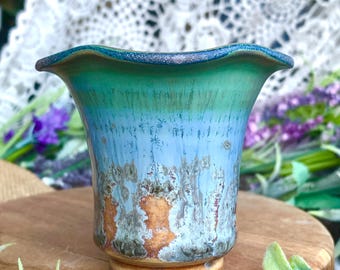 May include: A handcrafted ceramic vase with a flared rim and a pedestal base. The vase features a blend of turquoise, blue, and brown glaze, creating a unique, textured appearance. The vase is displayed on a wooden surface.