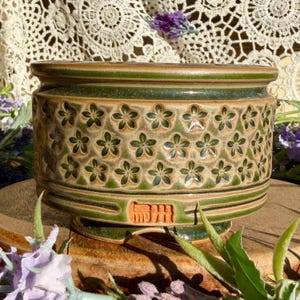 May include: A cylindrical ceramic planter featuring a repeating embossed floral pattern in dark green and tan. The rim and base are finished in a glossy dark green glaze. A rectangular stamp with Japanese characters is visible on the side.