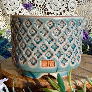 May include: A cylindrical ceramic planter featuring a teal and blue glaze with a repeating embossed floral pattern. The surface displays a grid of four-petaled flowers with light centers. A rectangular stamp with embossed characters is visible.