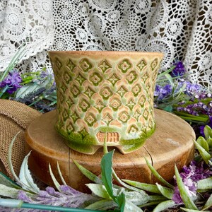 May include: A ceramic planter featuring a repeating geometric pattern of green and tan quatrefoil shapes. The pot has a flared rim, a tapered body, and a small stamped maker's mark near the base. The glaze has a subtle crackle texture.