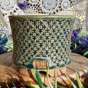 May include: A cylindrical ceramic planter featuring a deep green and blue glaze with a raised, light-colored lattice pattern of repeating floral and geometric shapes. The base is unglazed, textured clay with a rectangular stamp mark on the front.