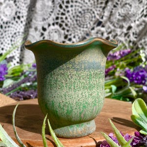 May include: A small, green ceramic vase with a speckled texture and a ruffled rim. The vase has a pedestal base and a slightly flared body. The rim is a darker shade of green. The vase is surrounded by purple flowers and greenery.