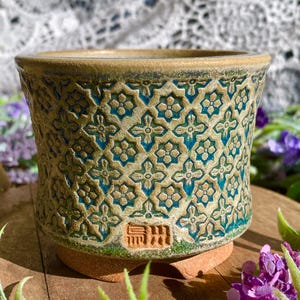 May include: A ceramic planter with a detailed floral pattern in teal and beige. The pot has a textured surface and a rounded shape. The base is a reddish-brown color, and the rim is a darker shade of beige. The planter is suitable for small plants.