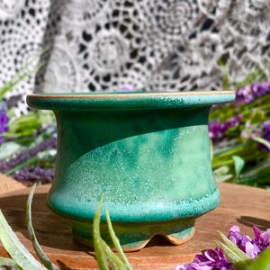 May include: A handcrafted, small, green ceramic planter with a speckled texture. The planter has a wide rim and a slightly flared body, sitting on a small base. The background includes purple flowers and a lace doily.
