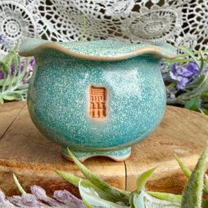May include: A small, round ceramic planter with a teal, speckled glaze and a wavy, flared rim. The pot features three short feet and a rectangular, unglazed terracotta-colored stamp on the front with embossed Asian characters.