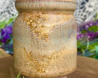 May include: A small, handcrafted ceramic jar with a textured, crystalline pattern in shades of gold, tan, and blue. The jar has a rounded body and a slightly flared rim, sitting on a small, pink base. The background is blurred.