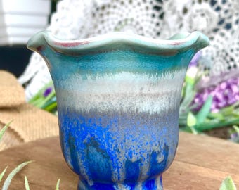 May include: A small, handcrafted ceramic vase with a ruffled rim. The vase features a gradient of colors, transitioning from a deep blue base to shades of gray and turquoise at the top. The rim has a touch of red.