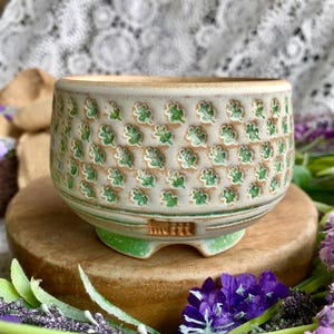 May include: A beige ceramic planter with a repeating pattern of green leaves. The planter has a rounded shape and sits on a small, green base. The planter is on a wooden surface.