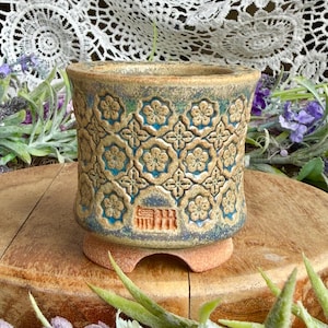 May include: A cylindrical ceramic planter featuring a repeating embossed floral pattern in tan against a blue glazed background. The base is unglazed terracotta with a cutout design, and a rectangular maker's mark is stamped on the side.