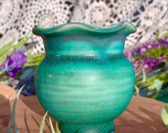 May include: A small, handcrafted ceramic vase with a vibrant teal glaze. The vase has a rounded body, a ruffled rim, and a subtle, textured surface. The vase is displayed on a wooden surface, surrounded by greenery and purple flowers.