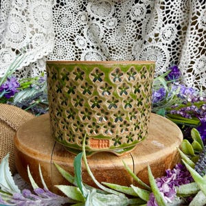 May include: A cylindrical ceramic planter featuring a repeating embossed floral pattern in shades of green and tan. The exterior surface displays a textured finish with a small rectangular stamp near the base containing four distinct characters.