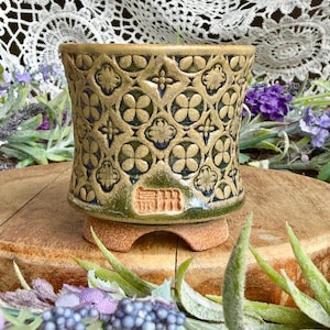 May include: A cylindrical ceramic planter with a tan and green glaze featuring a repeating embossed quatrefoil pattern. The base is unglazed terracotta with four small feet. A rectangular stamp with Japanese characters is visible on the front.