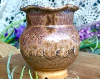 May include: A small, handcrafted ceramic vase with a unique, textured surface. The vase features a rounded body and a ruffled rim, with a warm brown and tan color palette. It sits on a small, light-colored base, perfect for displaying small flowers or decorative items.