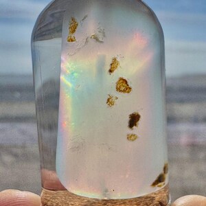 May include: A clear, glass bottle with a cork base. The bottle contains a light-colored, iridescent substance with brown inclusions. The bottle is cylindrical with a rounded top. The background is a blurred view of the sky and water.