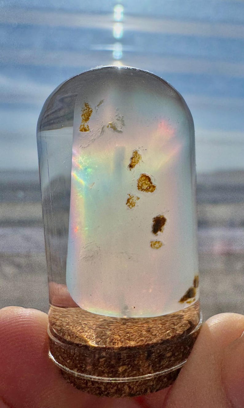 May include: A clear, glass bottle with a cork base. The bottle contains a light-colored, iridescent substance with brown inclusions. The bottle is cylindrical with a rounded top. The background is a blurred view of the sky and water.